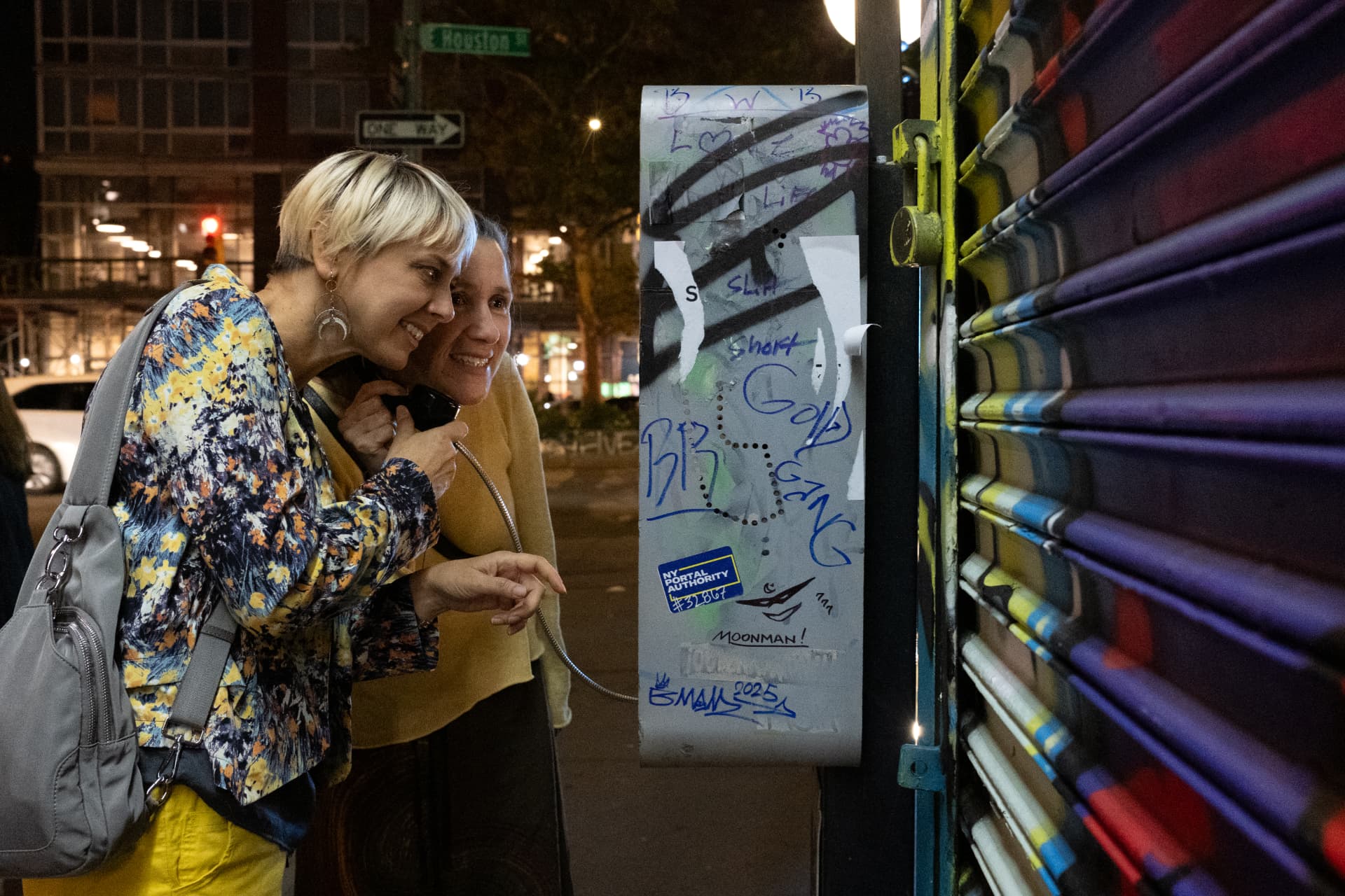 Two strangers sharing a payphone handset on a New York street at night, laughing